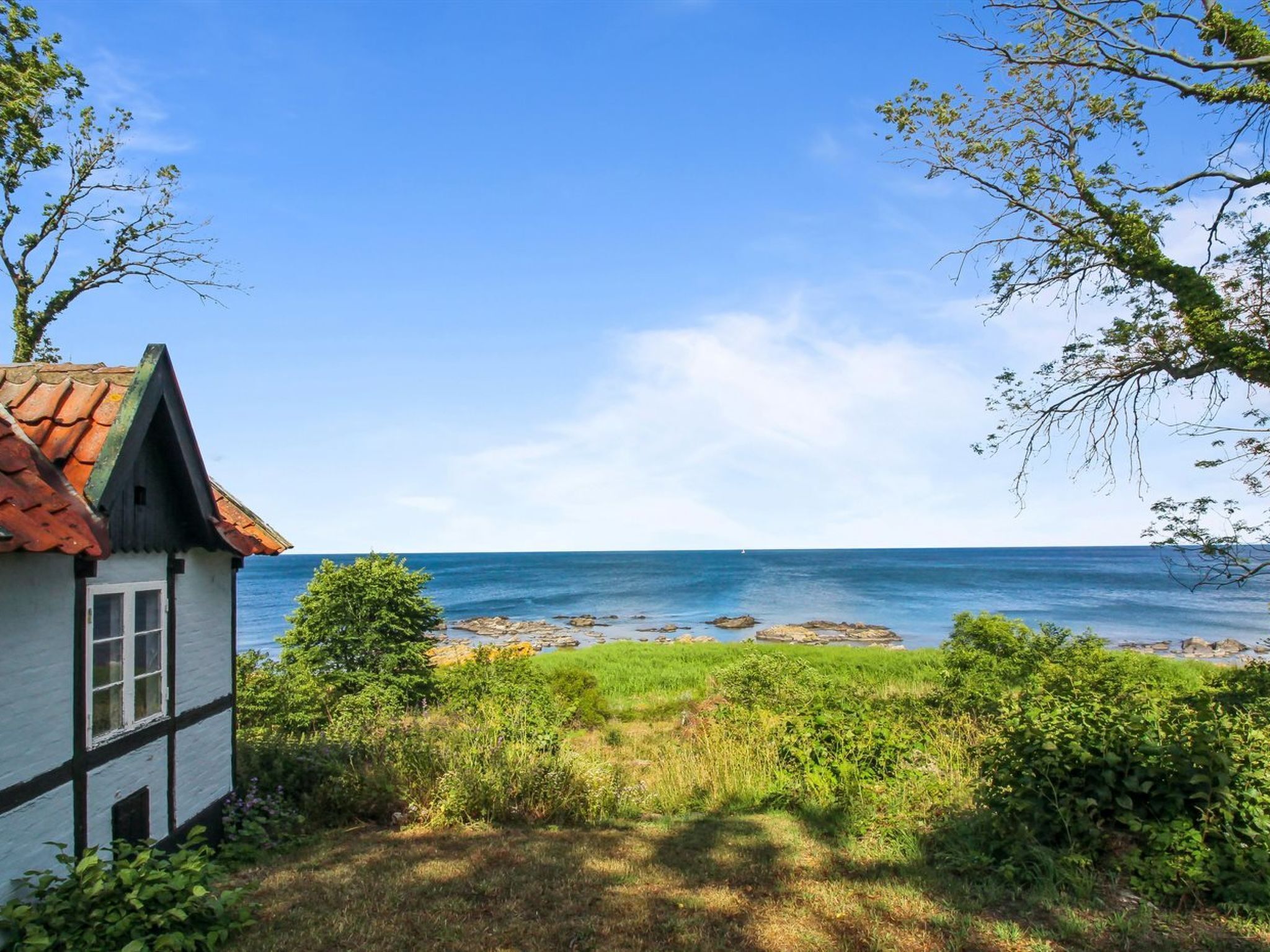 Uma casa costeira com fachada branca e telhado vermelho, localizada à beira-mar, com afloramentos rochosos na água.