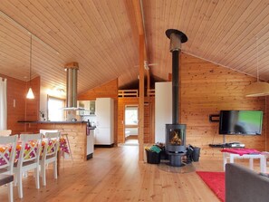 En mysig stuga med en central vedeldad kamin, en matplats med stolar och ett bord samt ett kök med kylskåp och skåp.