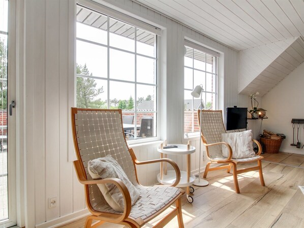 A room with two wooden chairs with mesh backs, a small round table, a TV on a stand, and large windows offering a view of the outside.