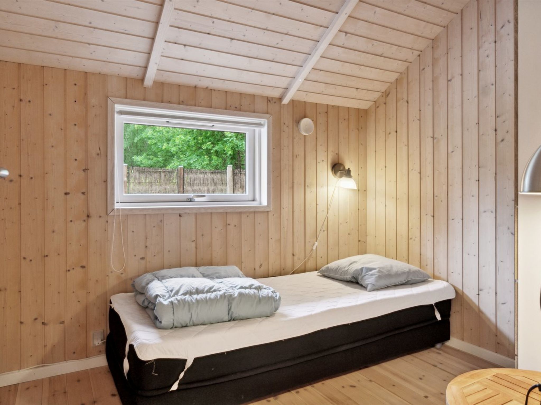 Une petite chambre avec un lit simple, des murs en bois, une fenêtre et une lampe fixée au mur.
