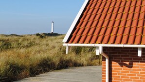 Un bâtiment avec un toit recouvert de tuiles rouges et un mur en briques, qui surplombe une zone herbeuse avec un phare visible au loin.