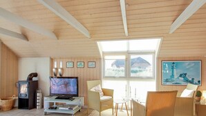 Un salon chaleureux avec un plafond en bois, une cheminée, un téléviseur, des meubles en osier et un tableau représentant un phare.