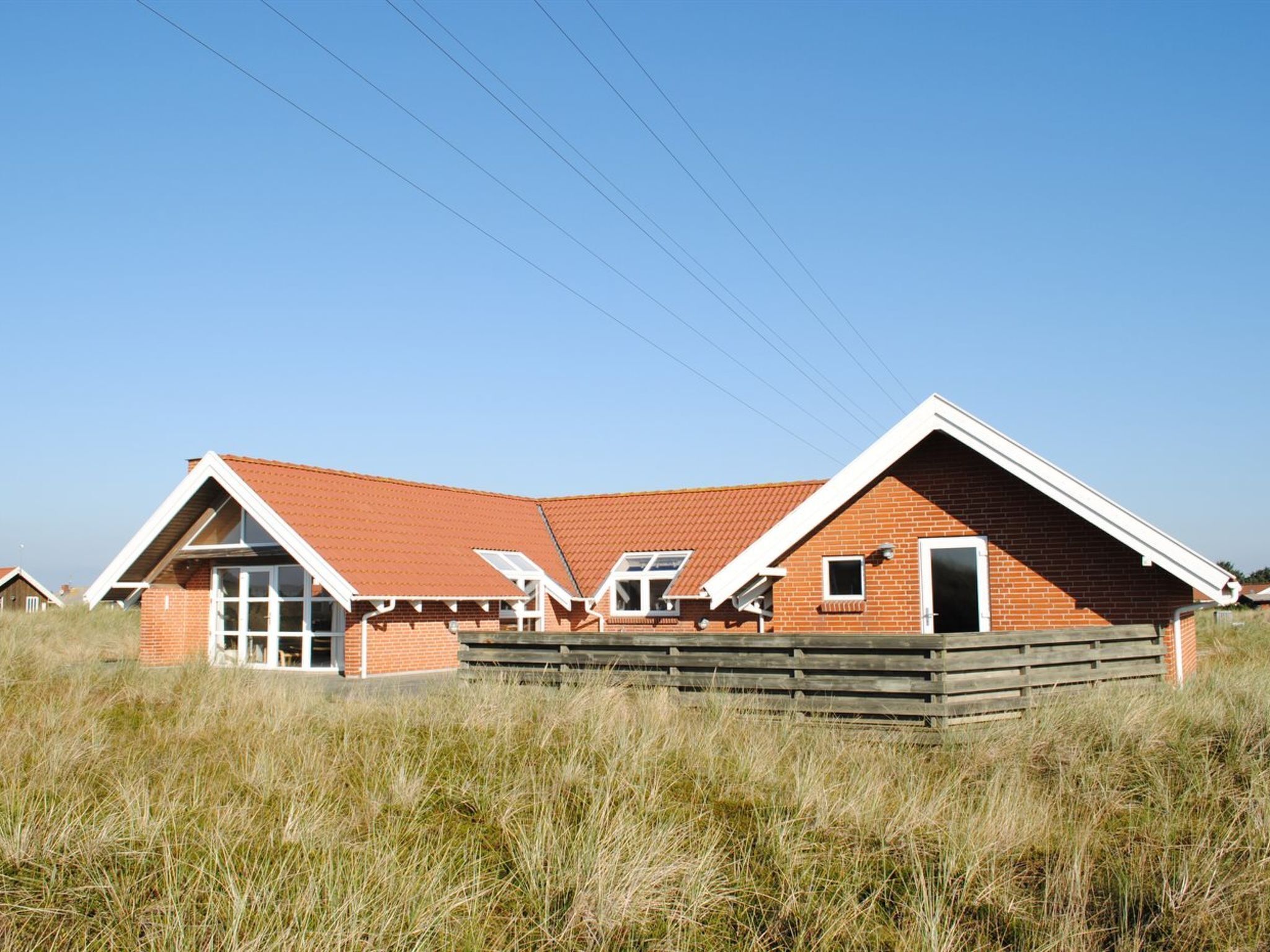 A two-story brick house with a sloped roof, large windows, and a wooden fence in a grassy field.