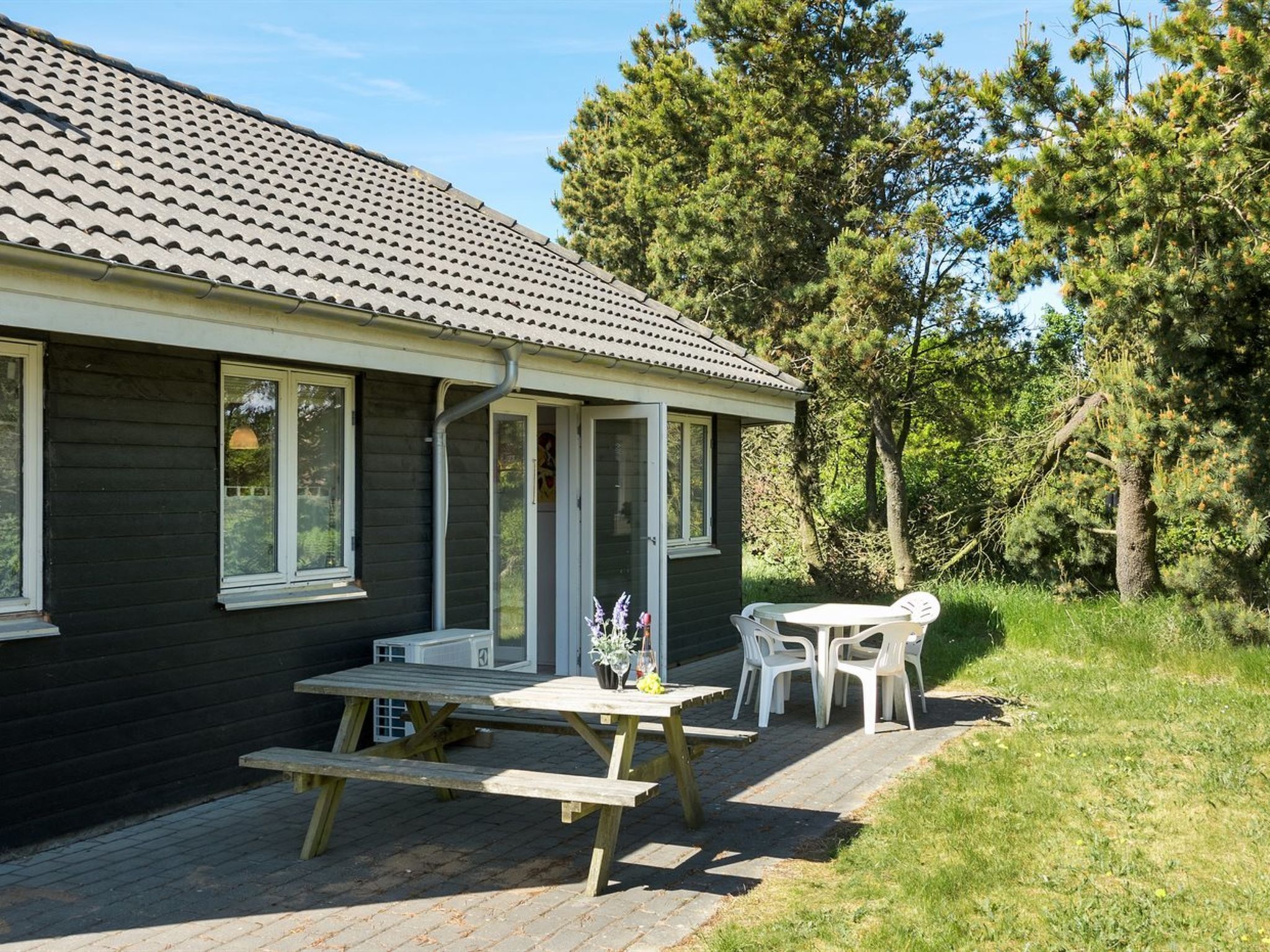 A wooden house with a tiled roof, a covered patio with a table and chairs, and a surrounding wooded area.
