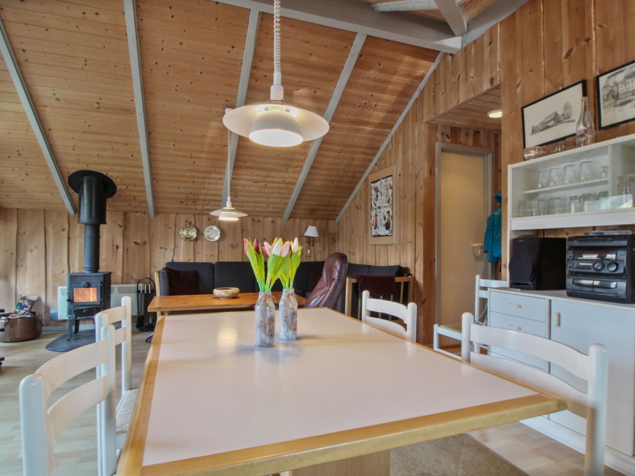 A cozy cabin interior with a wooden interior, a dining table with chairs, a sofa, a fireplace, and a shelf with glassware.