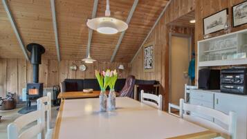 A cozy cabin interior with a wooden interior, a dining table with chairs, a sofa, a fireplace, and a shelf with glassware.