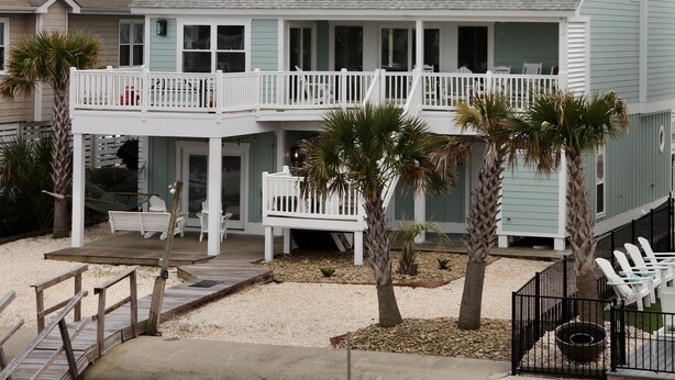 A two-story beach house with a balcony, outdoor seating, and palm trees.
