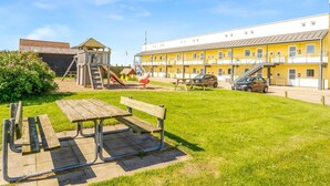 Een speeltuin met een glijbaan en klimconstructie, een picknicktafel, en een gebouw met meerdere balkons en een auto ervoor geparkeerd.