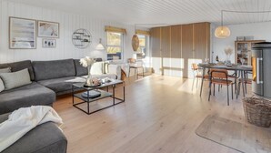 Ein modernes Wohnzimmer mit einem grauen Sofa, einem Couchtisch und einem Essbereich mit Tisch und Stühlen. Ein Kamin und ein Fenster mit Aussicht sind ebenfalls vorhanden.