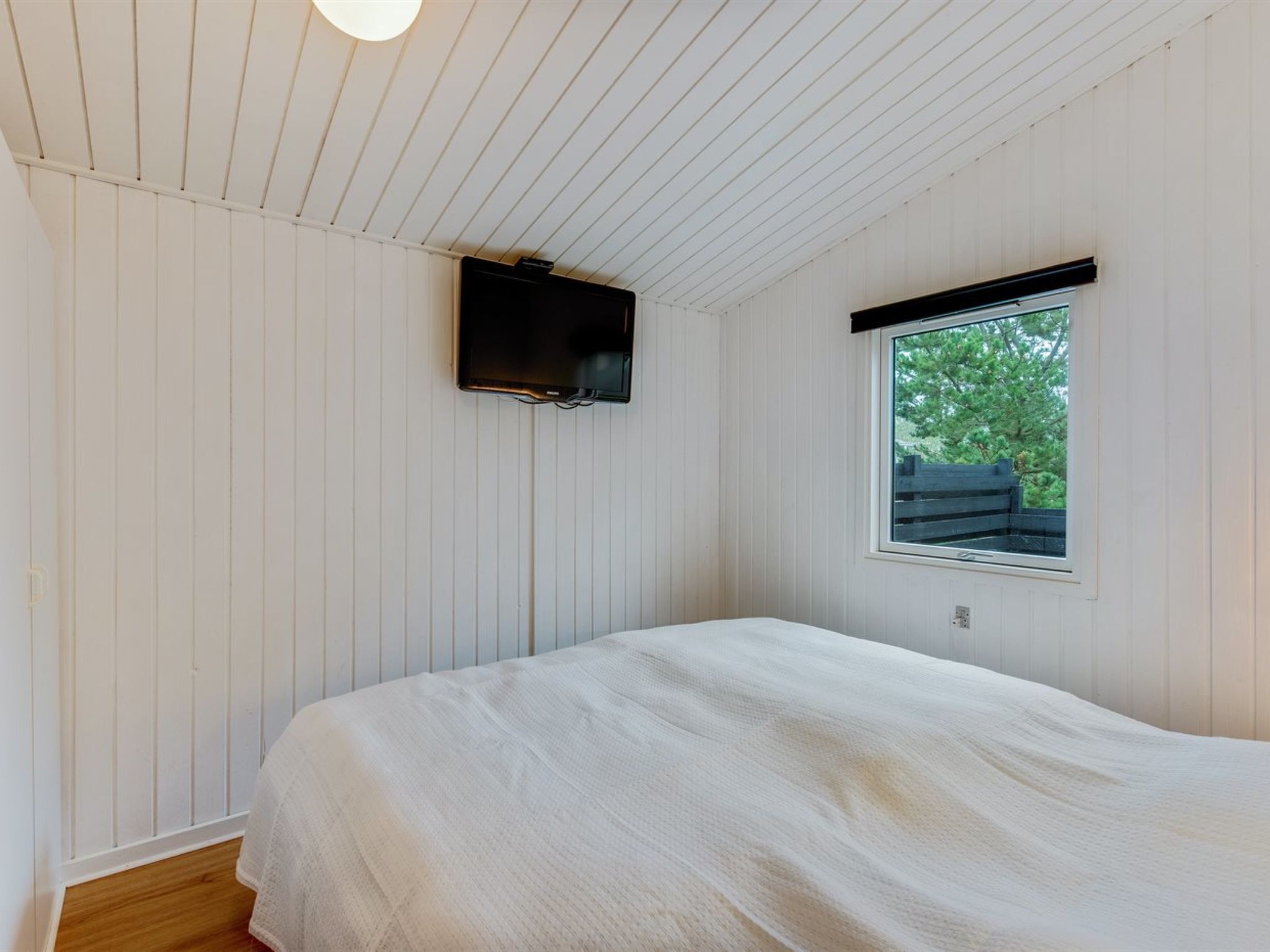 Quarto com cama, televisão fixada na parede e janela com vista para vegetação.