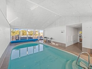 An indoor swimming pool with clear glass walls, a blue deck, and a seating area with chairs and a table.