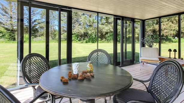 A covered patio with a round table and chairs, large glass windows, and a view of a grassy area. - "Biergh" - 600m from the sea by Interhome (Løkken)