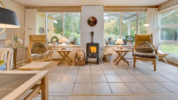 A cozy living room with a fireplace, wooden furniture, and large windows offering a view of the outdoors.