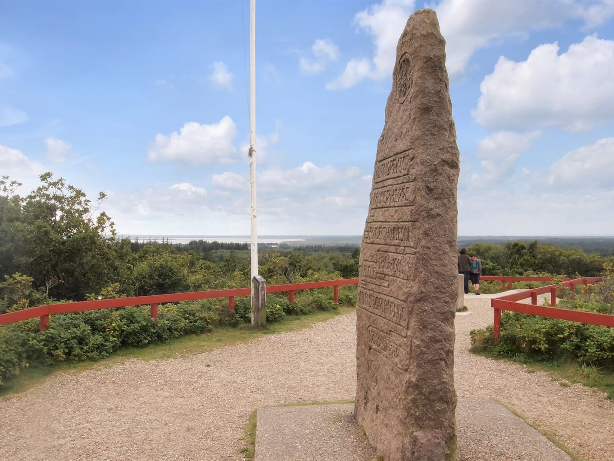 A tall stone monument with inscriptions, surrounded by a gravel path and red railings, overlooking a scenic landscape.