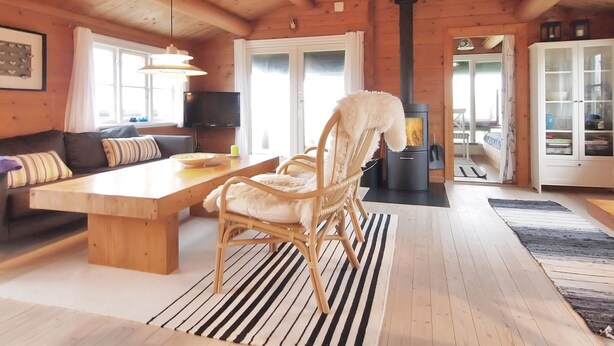 A cozy cabin interior with a wooden dining table, a sofa, a chair, a television, and a wood-burning stove.