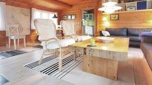 A cozy cabin interior with a wooden ceiling, a dining area with chairs and a table, a sofa, and a kitchen area.