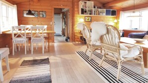 A cozy log cabin interior with a dining area, wicker chairs, a bookshelf, and a fireplace.