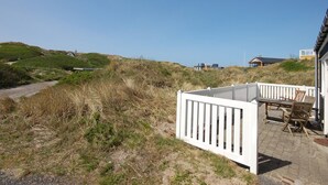 En fastighet vid kusten med ett vitt staket, utomhusmöbler och utsikt över stranden och sanddynerna.