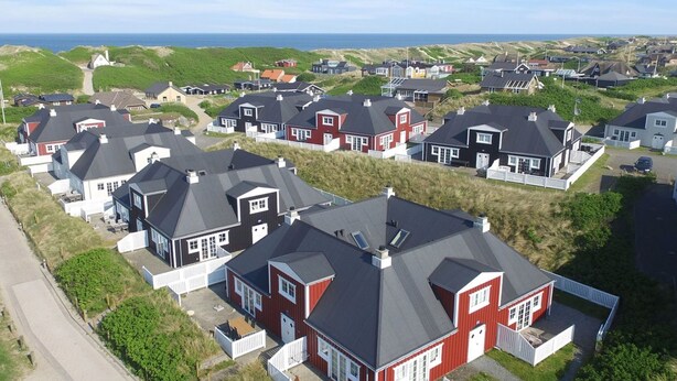 Ett bostadsområde vid kusten med hus som har mörka tak och röda detaljer, omgivet av gröna sanddyner och havet i bakgrunden.