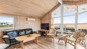 Una sala de estar acogedora con techo de madera, un sofá de cuero negro, una mesa de centro de madera, sillas de mimbre y grandes ventanales que ofrecen vistas al exterior.