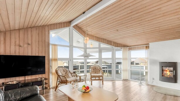 Amplia sala de estar con techo de madera, televisor grande, sofá de cuero negro, dos sillones, una mesa de centro de madera con fruta y una chimenea.