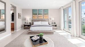 A modern bedroom with a large bed, two bedside tables, a nightstand, a window with curtains, and a view of the beach. - The Boardroom Residence at Peninsula Village (Newport Beach)