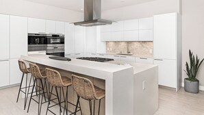 A modern kitchen with white cabinetry, a central island with bar stools, a stainless steel range, and a large overhead vent. - The Boardroom Residence at Peninsula Village (Newport Beach)