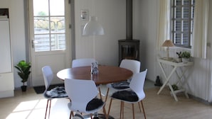 Een gezellig eetgedeelte met een ronde houten tafel, witte stoelen, een hangende lamp en een houtkachel.