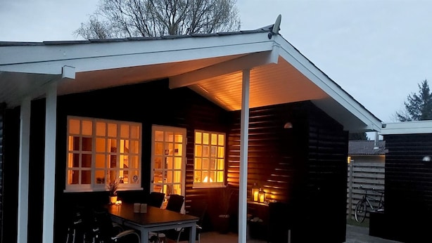 Een overdekt terras met een tafel en stoelen, een houten hut met ramen, en een fiets op de achtergrond.