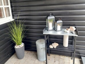 A black wooden exterior wall, a metal table with two lanterns and candles, a potted plant, and a metal trash can.