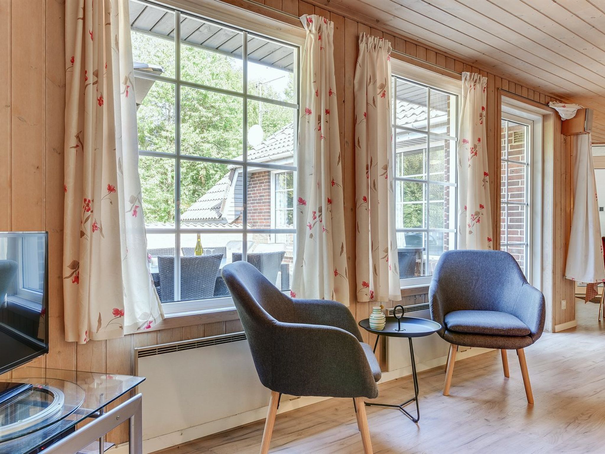 Een gezellige kamer met houten wanden, een televisie, twee fauteuils, een klein tafeltje en grote ramen met bloemen gordijnen.