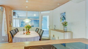 A modern dining area with a wooden ceiling, large windows, a round table with chairs, a cabinet with decorative items, and a kitchen area with a stove. - "Friederk" - 12km from the sea by Interhome (Oksbøl)