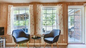 A room with wooden walls, two blue chairs, a small table with decorative items, and large windows with floral curtains. - "Friederk" - 12km from the sea by Interhome (Oksbøl)