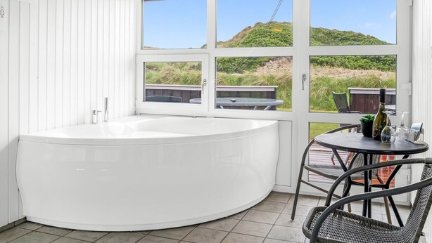 Ein modernes Badezimmer mit einer großen Badewanne, einem kleinen Tisch mit Stühlen und Blick auf den Strand durch große Fenster.