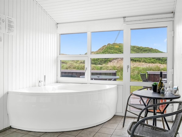 A modern bathroom with a large bathtub, a small table with chairs, and a view of the beach through large windows.