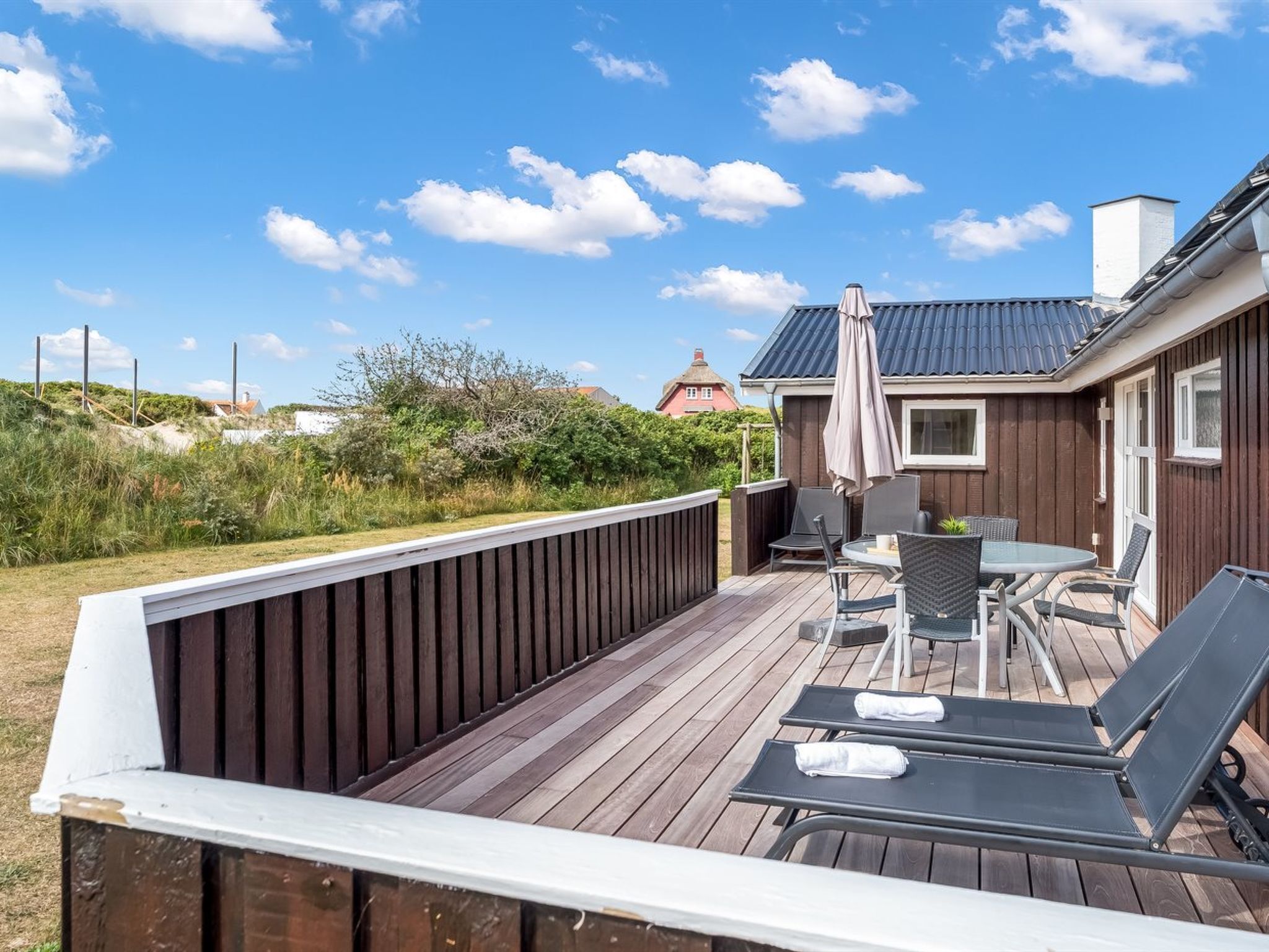 Una terraza de madera con una mesa y sillas, un espacio de patio cubierto y vista a una casa al fondo.
