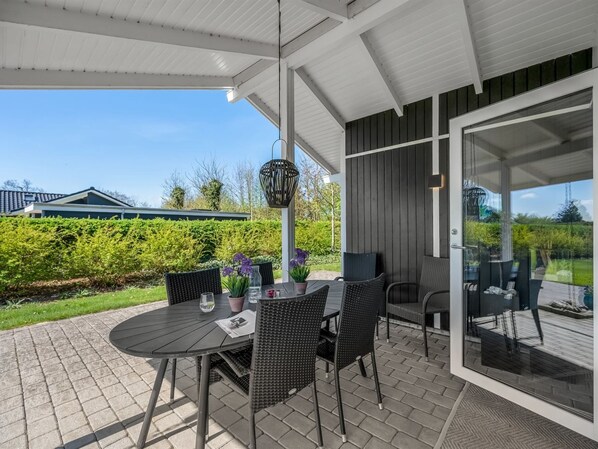 Une terrasse couverte avec une table et des chaises, un luminaire suspendu, et une vue sur un jardin et des bâtiments en arrière-plan.