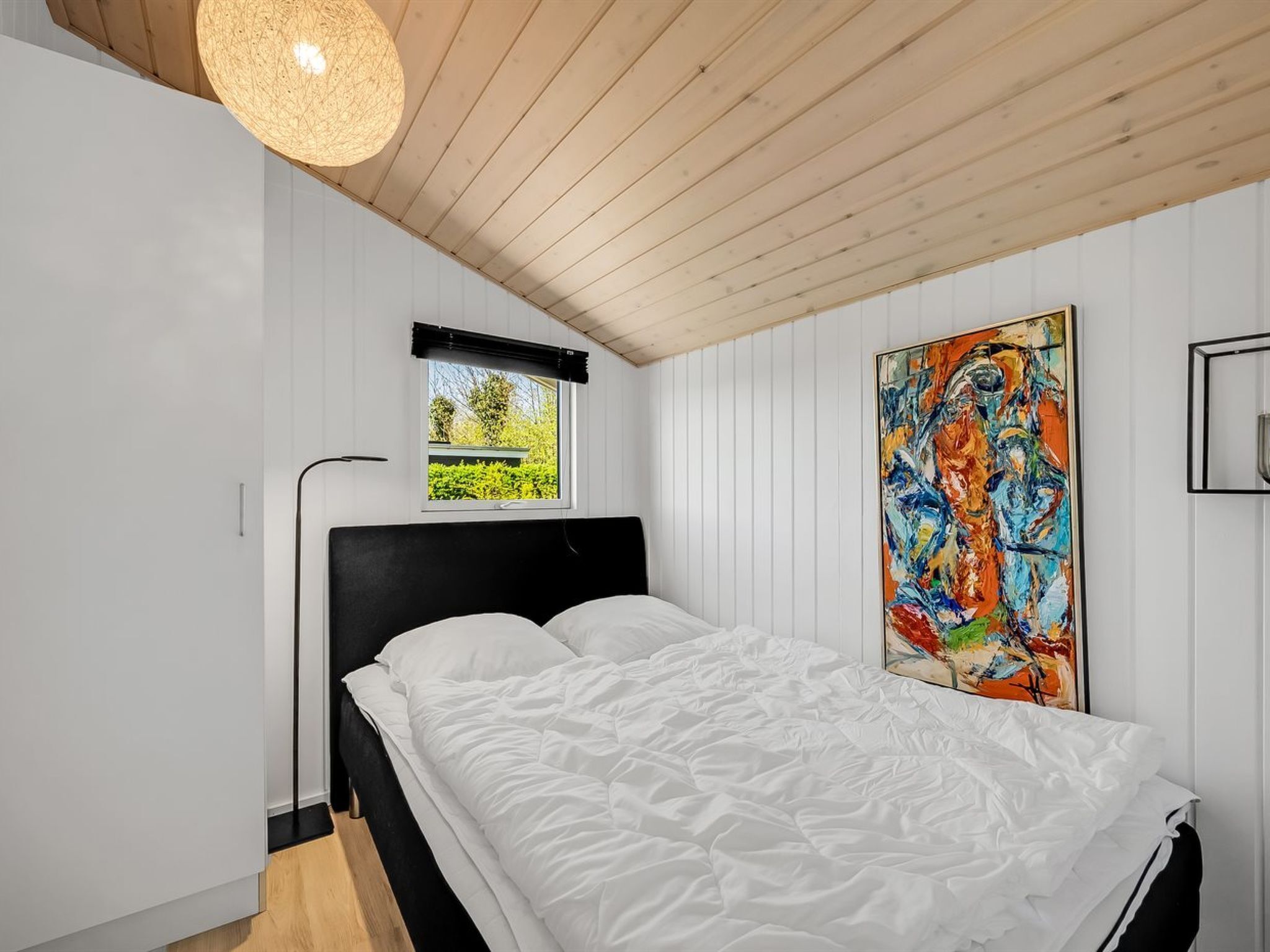 A bedroom with a bed, a window, a painting, and a ceiling light.