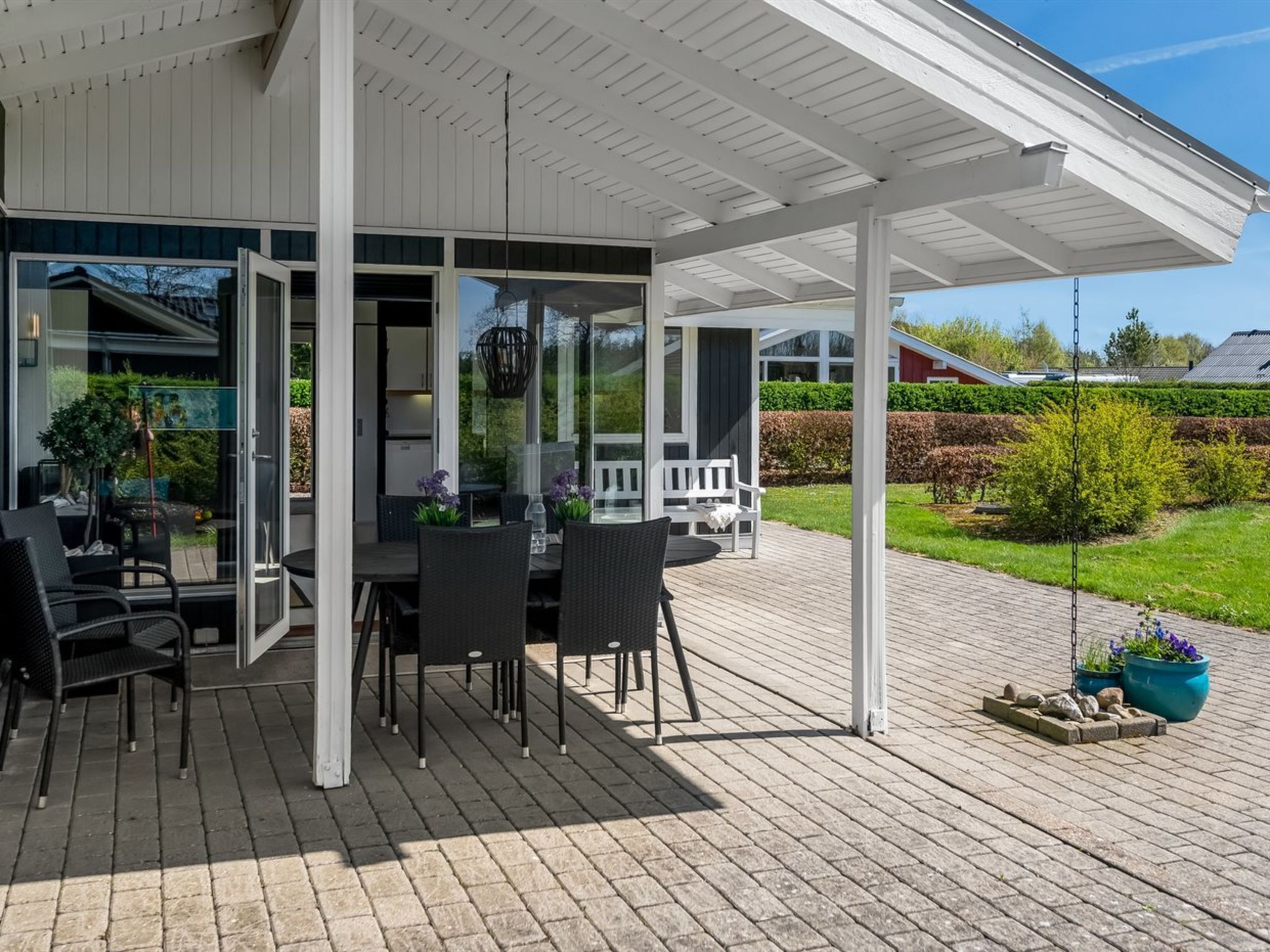 Eine überdachte Terrasse mit Tisch und Stühlen, eine Glastür zu einem Haus und ein Garten mit Bank und Blumentöpfen.