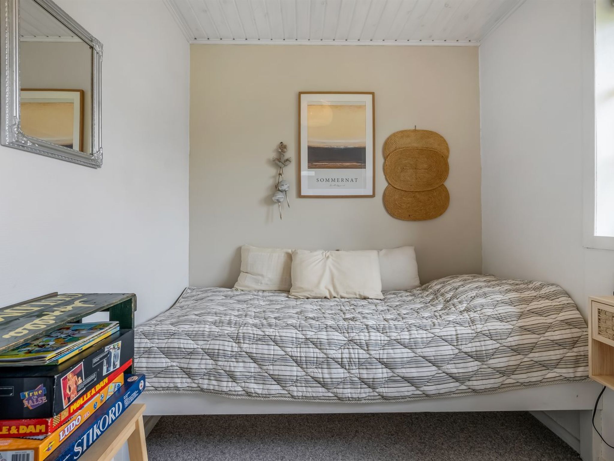 Quarto com cama, pilha de jogos de tabuleiro, quadro emoldurado e cesta de fibra.