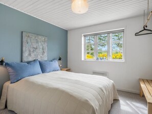 A bedroom with a bed, a window with a view of yellow flowers, a hanging light fixture, and a painting on the wall.