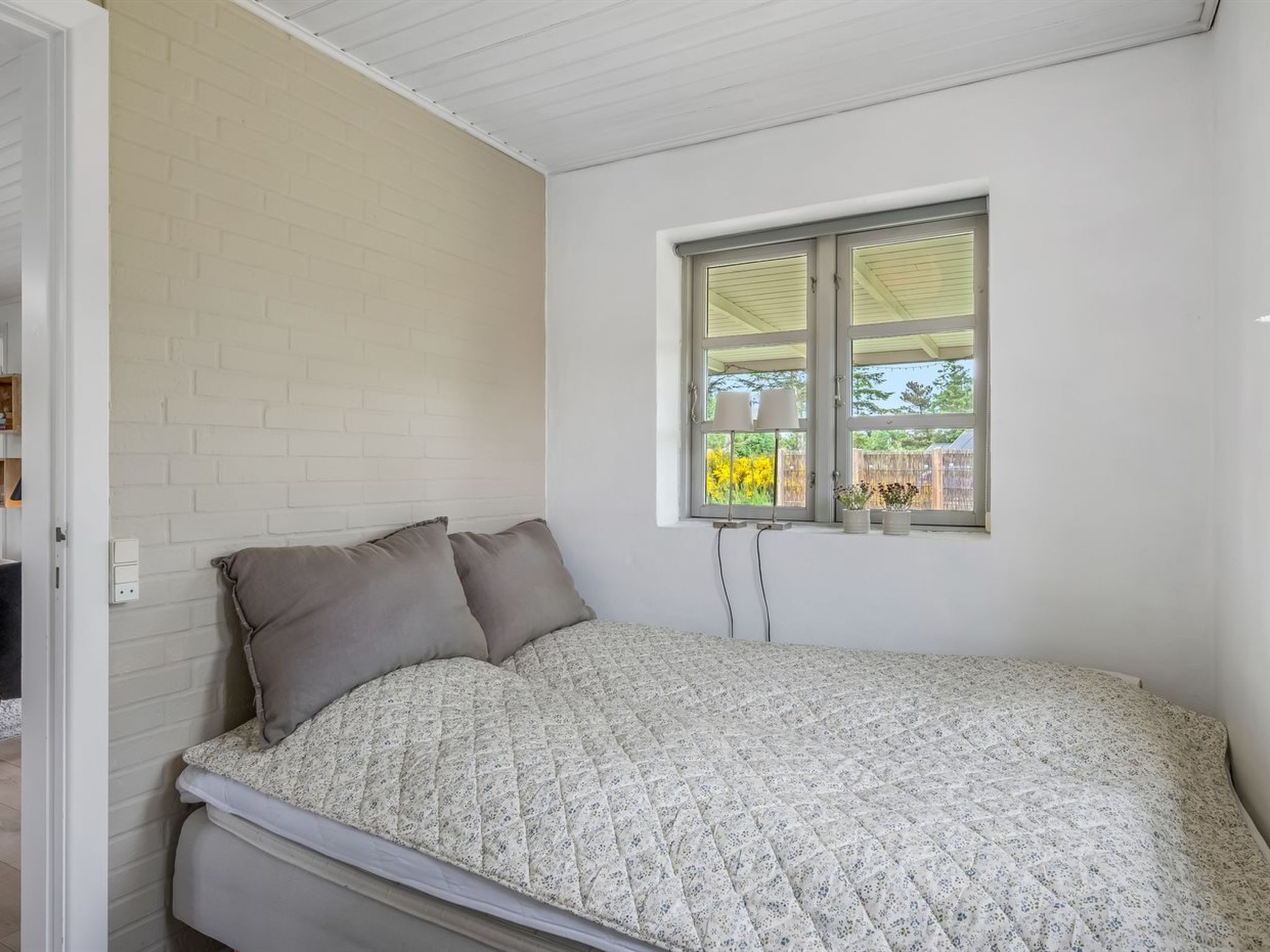 Quarto com cama, janela com vista para o exterior e parede de tijolos brancos.