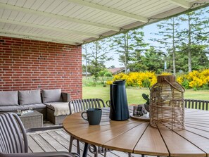 A covered patio with a round wooden table, wicker chairs, a cushioned sofa, and a brick wall.