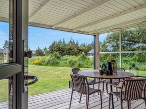 A covered outdoor patio with a table and chairs, a view of a grassy area and trees, and a house in the background.