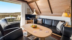 Un intérieur moderne avec un plafond en bois, un canapé en cuir noir, une table à manger en bois sur laquelle sont disposés des fruits et un jeu de cartes, et un téléviseur posé sur un meuble.