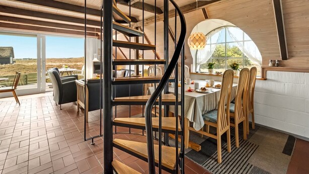 Un intérieur moderne avec un escalier en colimaçon, un coin repas avec des chaises en bois et une grande fenêtre donnant sur l’extérieur.
