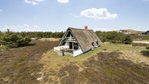 Une maison à toit de chaume, aux murs blancs, avec une cheminée bien visible et une petite terrasse agrémentée de chaises.