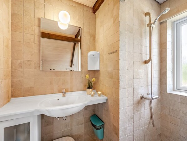 A bathroom with a sink, mirror, towel dispenser, shower, and a window.
