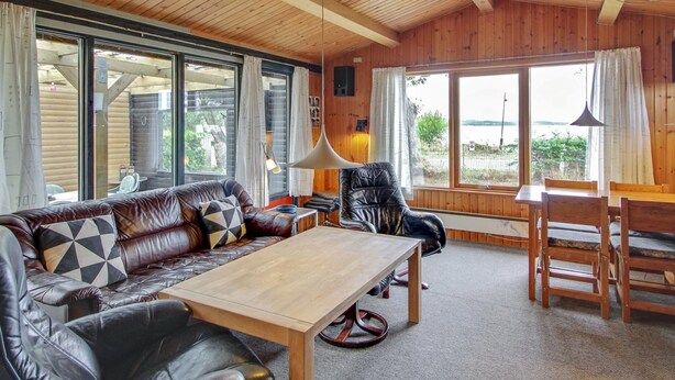 A cozy cabin interior with a wooden ceiling, leather sofa, a dining table, and large windows offering a view of the outdoors. - Vacation home "Graa" by Interhome (Skanderborg)