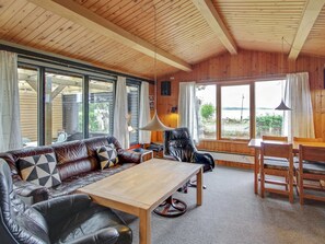 A cozy cabin interior with a wooden ceiling, leather sofa, a dining table, and large windows offering a view of the outdoors.